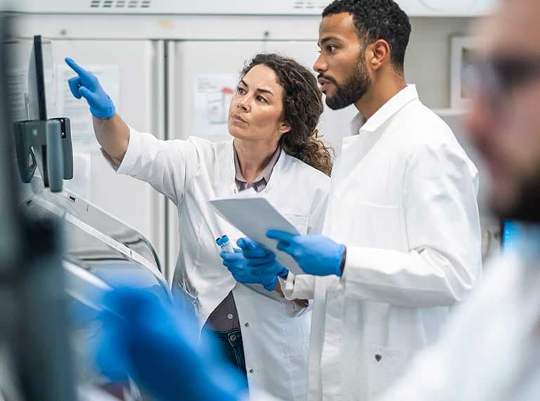 Two scientists in lab coats and blue gloves examining data on a monitor, one pointing at the screen.
