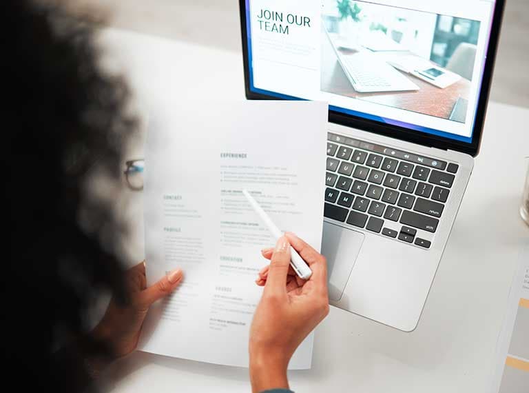Person reviewing CV next to laptop displaying job recruitment page with text "JOIN OUR TEAM" on screen