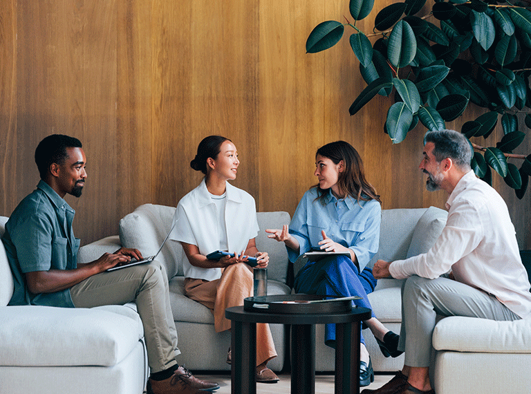 Four professionals sitting on white sofas having a discussion with notebooks against a wooden wall with green plant decor.
