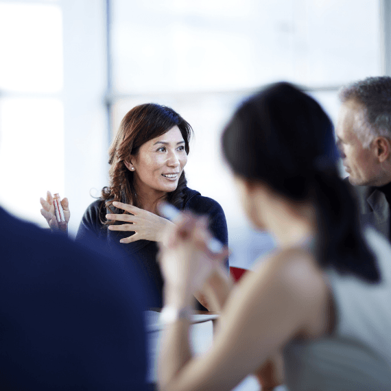 Woman with brown hair gesturing while speaking to a group of colleagues in a bright office meeting setting.