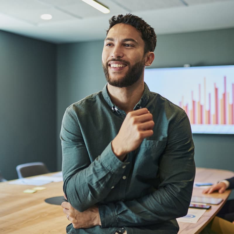 Confident young businessman standing in a modern office and smiling. He is wearing a casual shirt and has a beard. In the background, there are people sitting at a conference table.