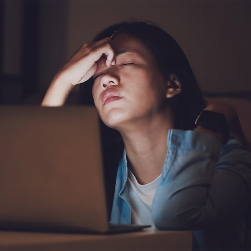 Woman with hand on forehead looking stressed or tired, illuminated by screen light in dim room wearing blue shirt.