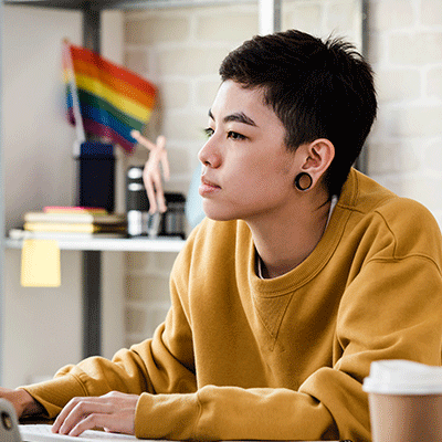 Person in yellow sweatshirt working at laptop with rainbow pride flag visible in background of modern office space.