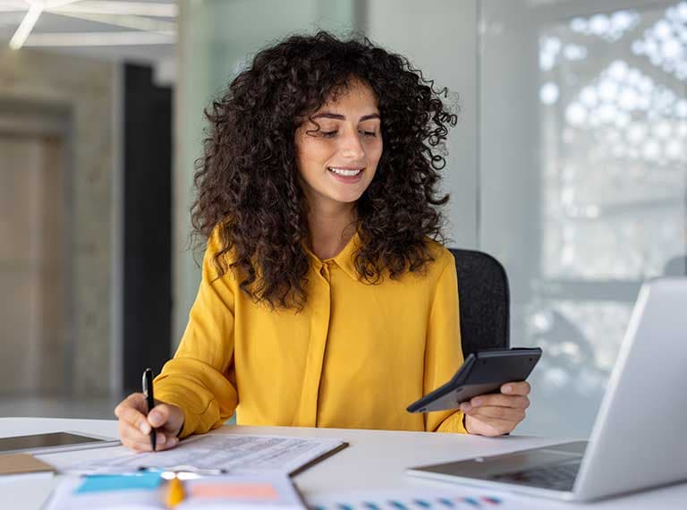 Person with curly hair in bright yellow blouse working at desk with laptop, phone, and documents in bright office.