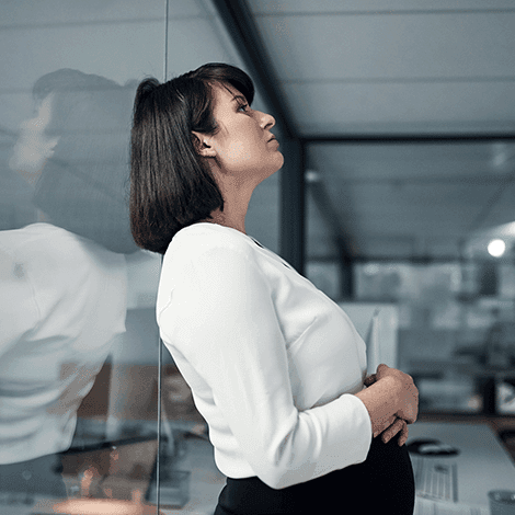 A white, slim, woman with a dark bob and fringe, wearing a white blouse and black skirt is standing against a glass, office wall, holding her stomach and staring upwards.