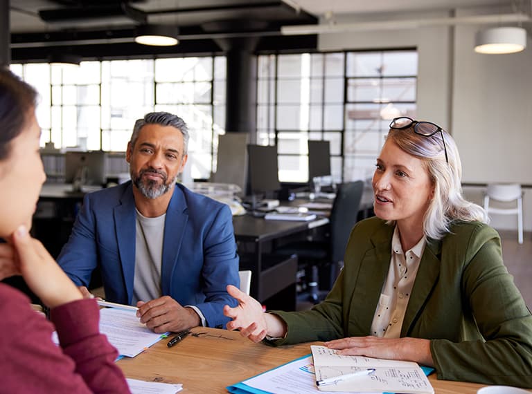 Three professionals in business meeting at wooden table in modern office with documents and computers in background.
