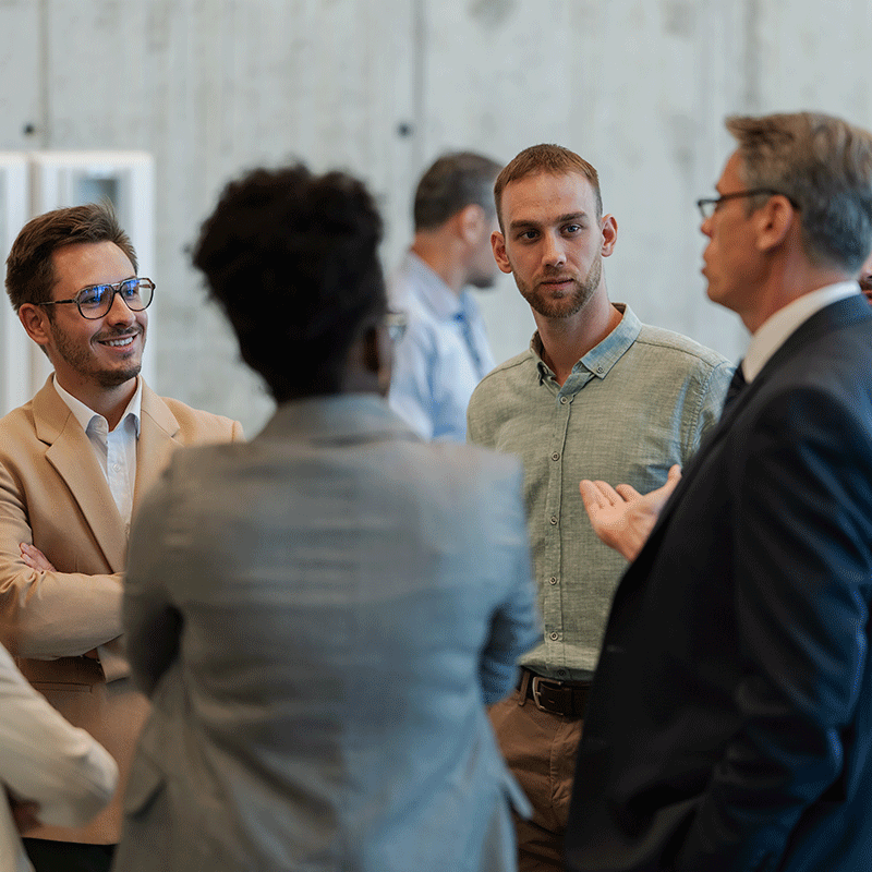 Business professionals in conversation at an office meeting, with a man in a tan blazer smiling and others engaged.