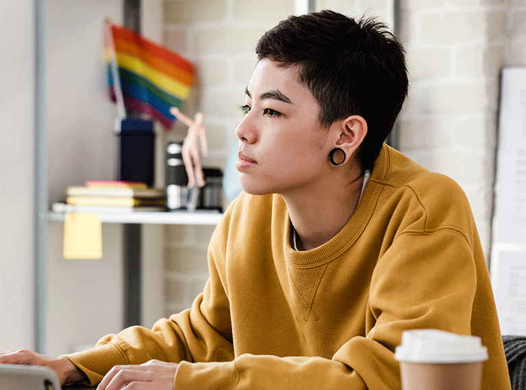 Person in mustard yellow sweatshirt sitting at desk with coffee cup, rainbow flag visible in background workspace.