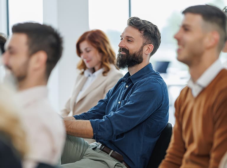 Bearded man in blue shirt smiling while sitting in a business meeting with colleagues in a bright office setting.