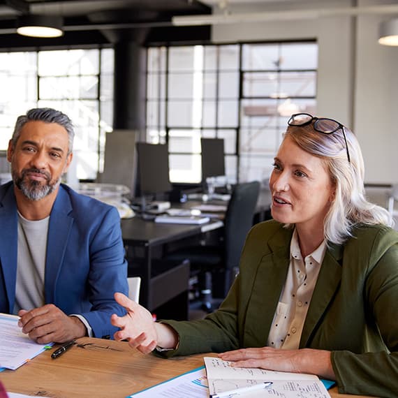 Two business professionals in discussion at a conference table in a modern office with glass walls and natural light.