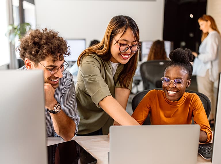 Three diverse coworkers smiling while collaborating at a laptop in a bright modern office setting.