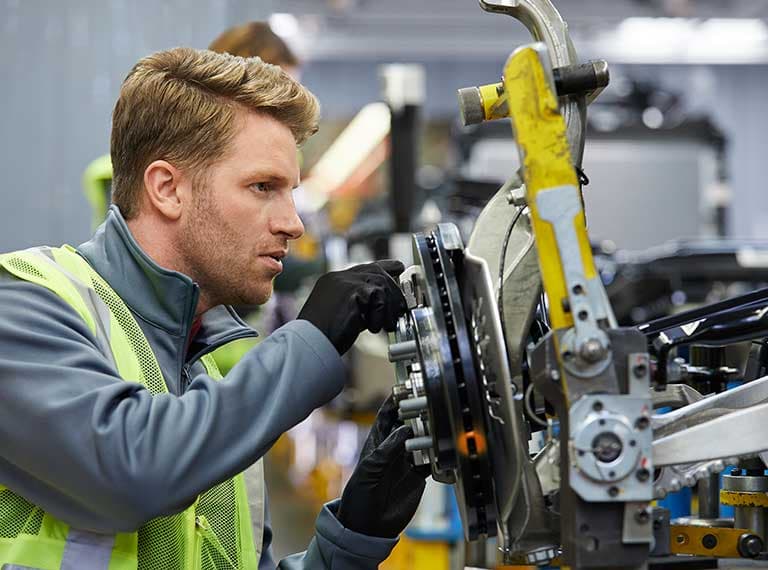 Automotive technician in safety vest examining a brake assembly on a vehicle in a manufacturing facility.
