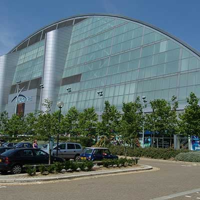 Modern curved glass building with metallic panels under blue sky, parking lot and green trees in foreground. Xscape MK.