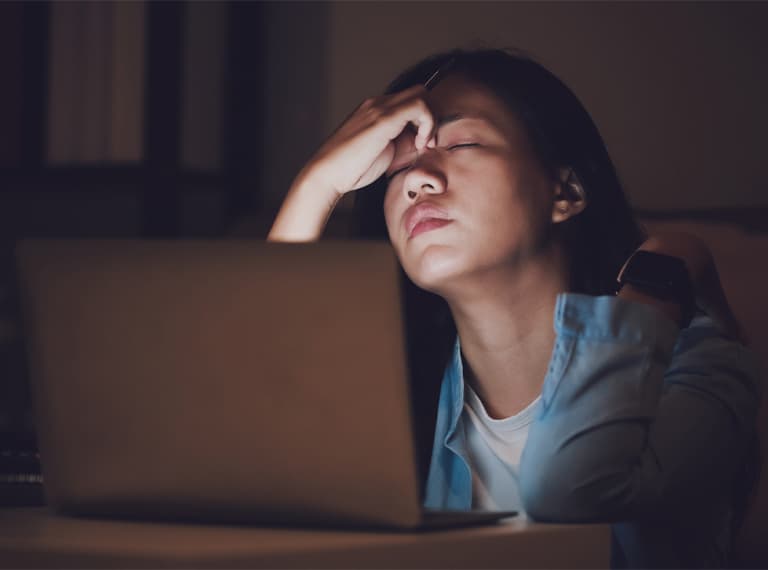 Woman with hand on forehead looking stressed while working on laptop in dim lighting, appearing tired or frustrated.