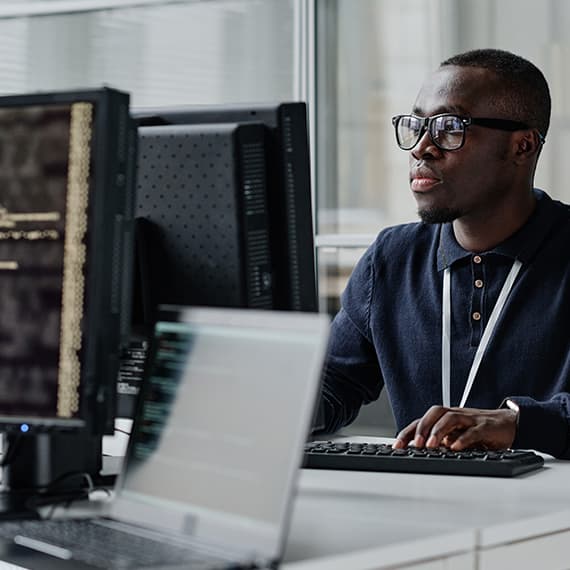 Man wearing glasses and navy blue polo shirt working at computer desk with dual monitors in modern office setting.