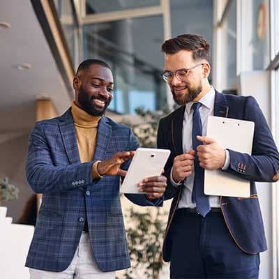 Two businessmen in suits reviewing information on a tablet and clipboard in a modern office building.