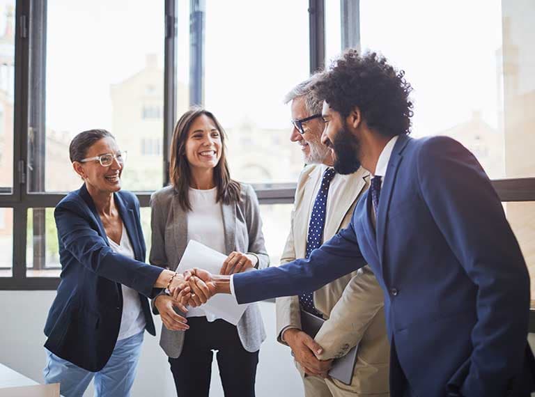 Four diverse business professionals smiling and shaking hands in a bright modern office with large windows.