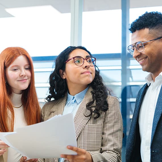 Three diverse young professionals reviewing a document together in a bright office with blue-framed windows in the background.