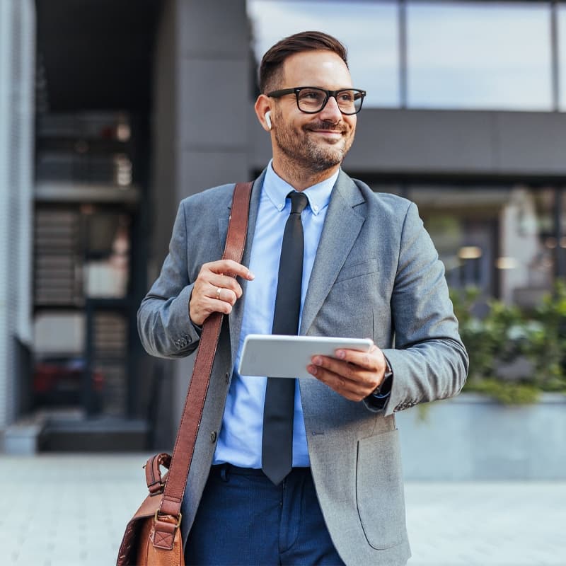 Smiling businessman in a suit and tie walking outside a modern office building, holding a tablet. He appears confident and professional, suggesting a successful business environment.