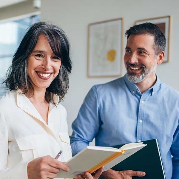 Smiling woman in white shirt and man in blue shirt holding notebook in bright office with framed artwork on wall