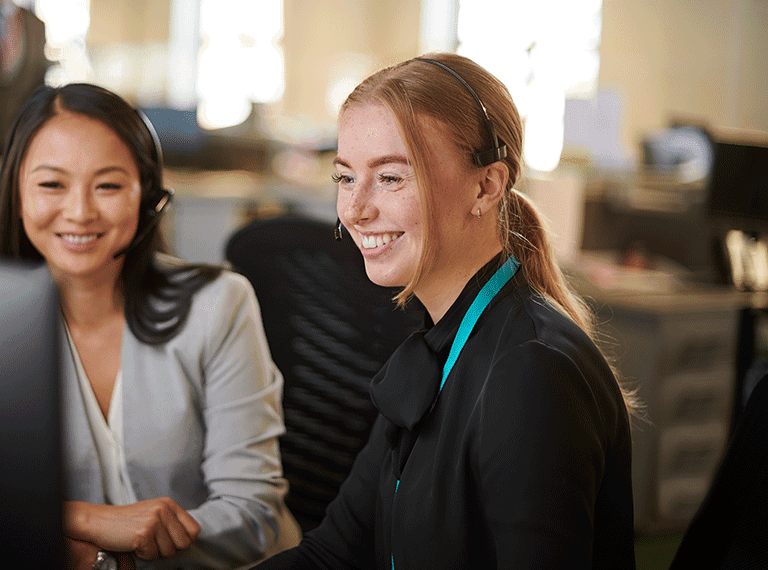 Two smiling professional women in an office setting, one wearing a black shirt with teal accent, the other in gray blazer.