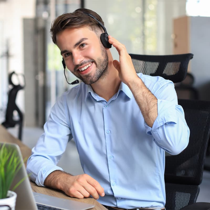Smiling male call-centre operator with headphones sitting at modern office, consulting online information in a laptop, looking up information in a file in order to be of assistance to the client.