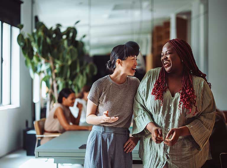 Two women laughing together in a bright modern office with colleagues working in the background near windows and plants.