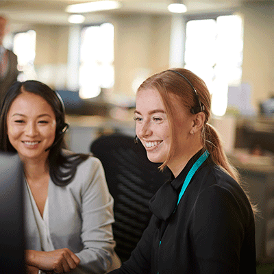 Three smiling professionals in an office setting, with a woman in black with turquoise accent in focus in the foreground.